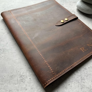 Può includere: Un diario in pelle marrone con chiusura a cinturino e ferramenta color ottone. Il diario presenta le iniziali "R.N.B." in rilievo nell'angolo in basso a destra. La pelle ha un aspetto invecchiato con cuciture visibili.