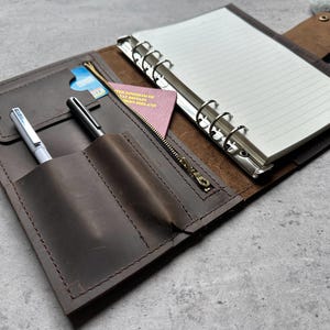 Könnte beinhalten: Braunes Lederjournal mit Stifthalter, Reißverschlusstasche und liniertem Papier. Das Journal hat einen silbernen Ordner und enthält einen Reisepass und eine Kreditkarte. Zwei Stifte befinden sich im Stifthalter.