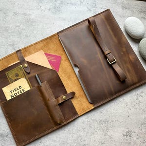 Puede incluir: Carpeta de cuero marrón abierta, con múltiples bolsillos y un portalápices. Una correa con hebilla de latón asegura la carpeta. En el interior, un bloc de notas con las palabras "FIELD NOTES" es visible, junto con un pasaporte y dinero en efectivo.