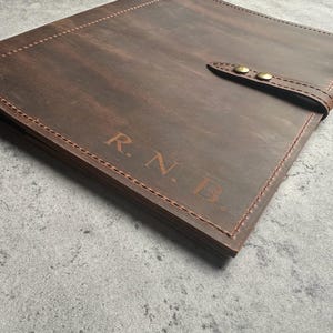 May include: A brown leather portfolio with the initials "R.N.B." embossed on the cover. The portfolio has a strap closure with two brass-colored buttons and is stitched with brown thread. The leather has a distressed appearance.
