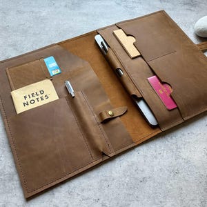 Op de afbeelding: Een bruin leren folio met meerdere vakken en compartimenten. Het heeft een penhouder, een klein notitieblok met de tekst "FIELD NOTES" en sleuven voor kaarten en een paspoort. De folio is beveiligd met een leren riem en een knoopsluiting.