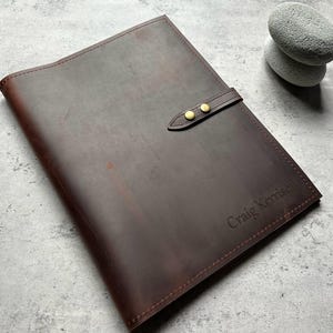 Puede incluir: Un portafolio de cuero marrón con cierre de correa y remaches dorados. El portafolio tiene el nombre "Craig Kerrison" grabado en la esquina inferior derecha. El cuero tiene un aspecto envejecido.