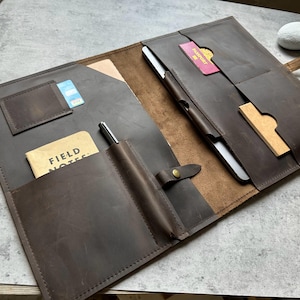 Puede incluir: Carpeta de cuero marrón abierta con múltiples bolsillos, que contiene un bolígrafo, un bloc de notas, una tarjeta de crédito y un pasaporte. La carpeta se asegura con una correa de cuero y un cierre de botón. El bloc de notas dice "FIELD NOTES".