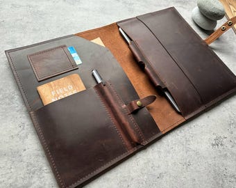 May include: Open brown leather portfolio with multiple pockets and pen holders. Includes a small card slot and a pocket holding a wooden Field Notes notebook. A silver pen is in a leather pen holder. The portfolio is secured with a leather strap.