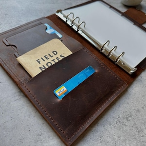 May include: Open brown leather binder with a card slot, holding a "FIELD NOTES" notepad, and a blue Visa card. The binder has a silver ring mechanism and a clear plastic sheet. The leather has a distressed look.