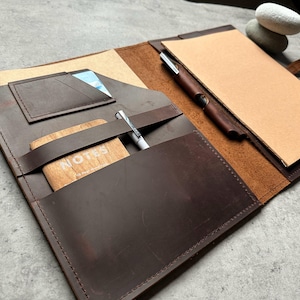 May include: Open, dark brown leather notebook with a notepad, pen, and card slots. A wooden card with the word "NOTES" is visible. The notebook is open, revealing a light brown interior and a pen holder.