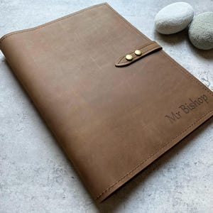 May include: A brown leather portfolio with a strap closure and two brass-colored rivets. The text "Mr Bishop" is embossed on the lower right corner. Two small, smooth stones are in the background.
