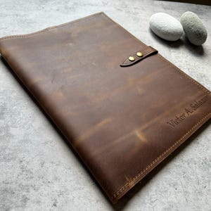 May include: A brown leather portfolio with a brass clasp closure. The portfolio has a personalized inscription that reads "Victor A. Salazar". The leather has a distressed finish.