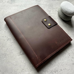 May include: A dark brown leather portfolio with a flap closure secured by two brass-colored rivets. The portfolio has a stitched border and the word "Piechota" is embossed on the lower right corner. Two smooth, gray stones rest in the background.