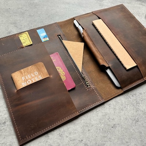 Puede incluir: Carpeta de cuero marrón abierta con múltiples bolsillos y compartimentos. Incluye un bolígrafo, un bloc de notas y una regla. Cuenta con ranuras para tarjetas y un bolsillo con cremallera. La carpeta está abierta, revelando su contenido.