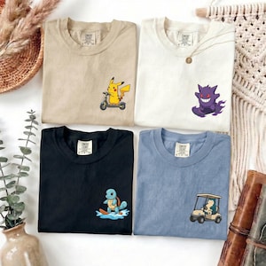 Può includere: Quattro magliette beige, bianche, nere e blu, ognuna con una grafica di un personaggio dei cartoni animati diverso. La maglietta beige ha Pikachu su uno scooter, quella bianca Gengar che fa surf, quella nera Squirtle che fa surf e quella blu Snorlax in un golf cart.