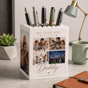 Könnte beinhalten: Ein weißer Stifthalter mit Blockdesign, mit Familienfotos und dem Text "WE LOVE YOU" und "Daddy". Der Halter enthält mehrere Stifte in verschiedenen Farben. Der Text "My Hero. My Guide. My Forever." ist ebenfalls sichtbar.