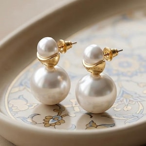 Puede incluir: Un par de pendientes de perlas. Cada pendiente presenta una gran perla blanca redonda en la base, coronada por una perla más pequeña y detalles metálicos dorados. Los pendientes se exhiben en una bandeja ovalada decorativa con un patrón floral.