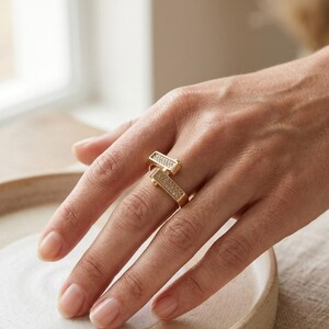 May include: A gold-colored ring with two rectangular sections, each encrusted with small, sparkling stones. The ring is worn on a finger, showcasing its modern, geometric design. The ring's design is a contemporary take on a classic jewelry piece.