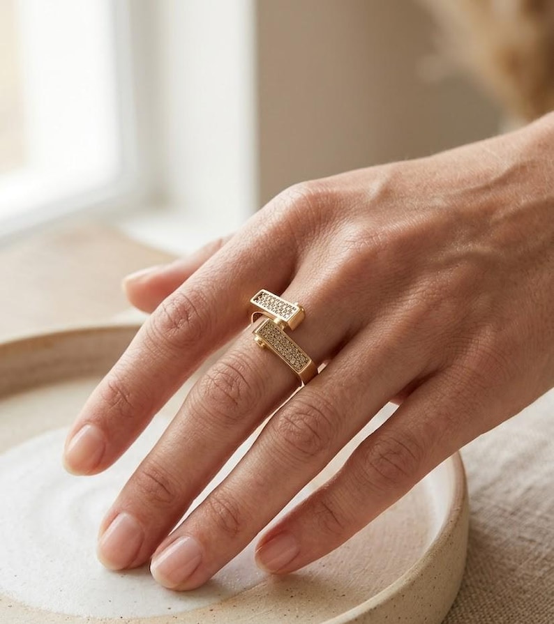 May include: A gold-colored ring with two rectangular sections, each encrusted with small, sparkling stones. The ring is worn on a finger, showcasing its modern, geometric design. The ring's design is a contemporary take on a classic jewelry piece.