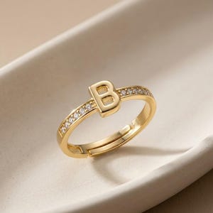 May include: A gold-coloured adjustable ring featuring the letter "B" in a serif font. The ring band is decorated with small, clear gemstones. The ring is displayed on a white, curved surface.