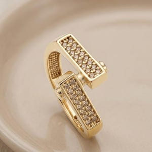 May include: A gold-colored, hinged clasp with two rectangular sections encrusted with small, sparkling stones. The clasp is set against a soft, neutral background, highlighting its intricate design and polished finish. The jewelry piece is likely used for necklaces or bracelets.