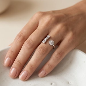 Puede incluir: Dos anillos de plata con piedras preciosas transparentes. Uno presenta un diseño en forma de lazo con piedras rectangulares, el otro un racimo floral de piedras redondas. Los anillos se muestran en una mano con uñas cuidadas.