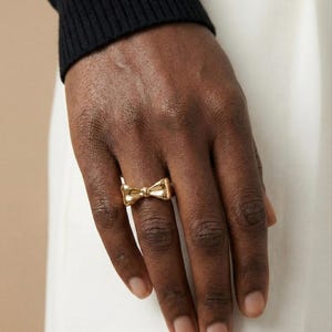 Può includere: Un anello dorato a forma di fiocco, indossato su una mano dalla pelle scura. Il design dell'anello è al centro dell'immagine, con la sua superficie lucida e riflettente. Lo sfondo include una manica di maglione nera e tessuto bianco.