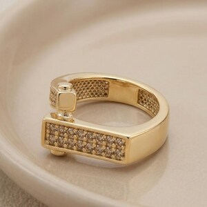 May include: A gold-colored ring with a unique design. The ring features a rectangular section encrusted with small, clear stones. The band has a textured pattern and a square accent. The ring is displayed on a light-colored surface.