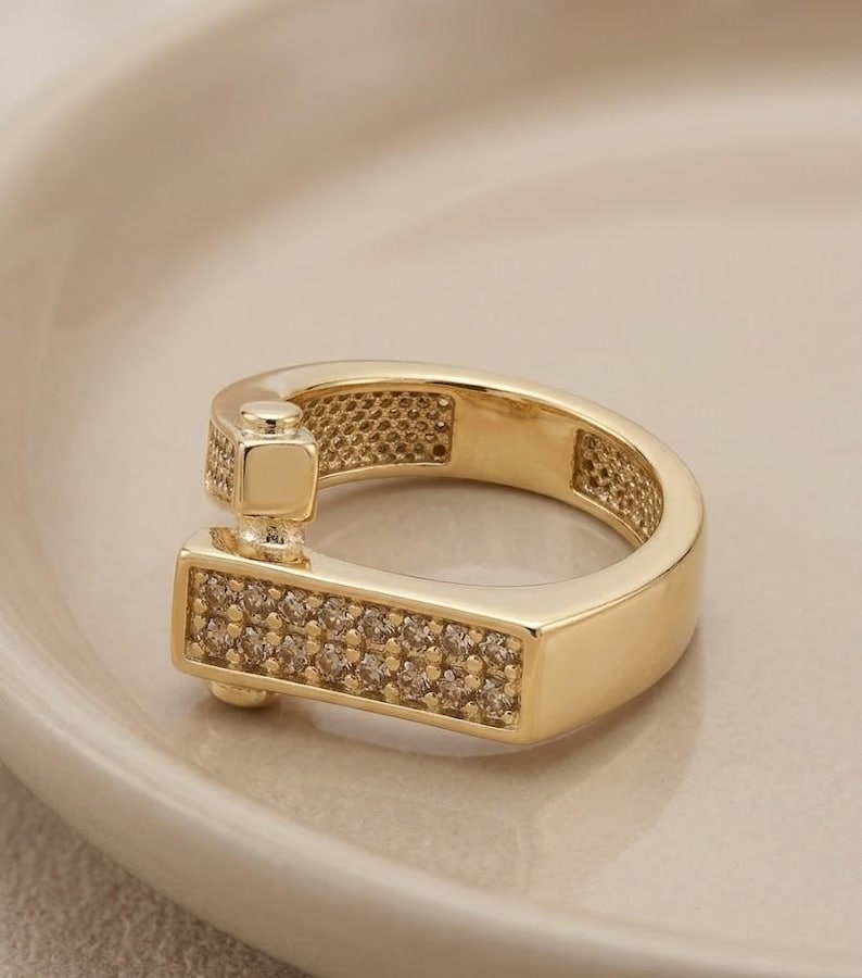 May include: A gold-colored ring with a unique design. The ring features a rectangular section encrusted with small, clear stones. The band has a textured pattern and a square accent. The ring is displayed on a light-colored surface.