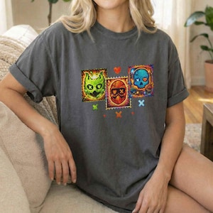 K&ouml;nnte beinhalten: Dunkelgraues T-Shirt mit drei farbenfrohen Sch&auml;delmotiven, die Briefmarken &auml;hneln. Die Designs zeigen ein gr&uuml;nes Monster, eine rot behelmte Figur und einen blauen Sch&auml;del. Kleine Herzen und Kreuze akzentuieren das Design.