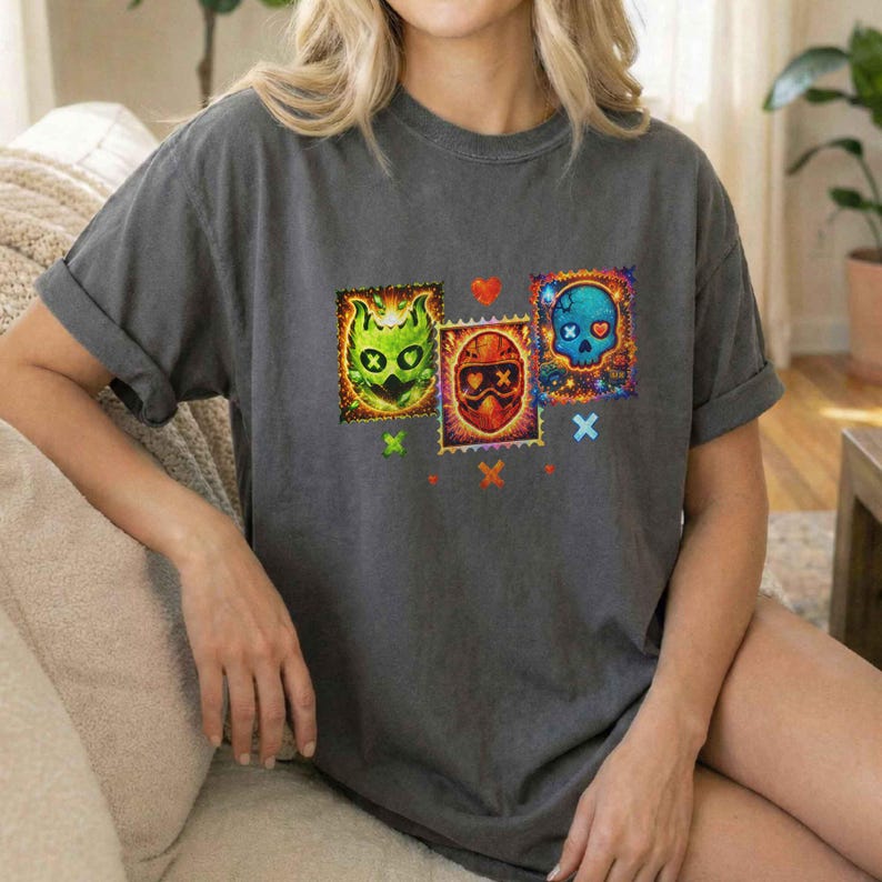 K&ouml;nnte beinhalten: Dunkelgraues T-Shirt mit drei farbenfrohen Sch&auml;delmotiven, die Briefmarken &auml;hneln. Die Designs zeigen ein gr&uuml;nes Monster, eine rot behelmte Figur und einen blauen Sch&auml;del. Kleine Herzen und Kreuze akzentuieren das Design.