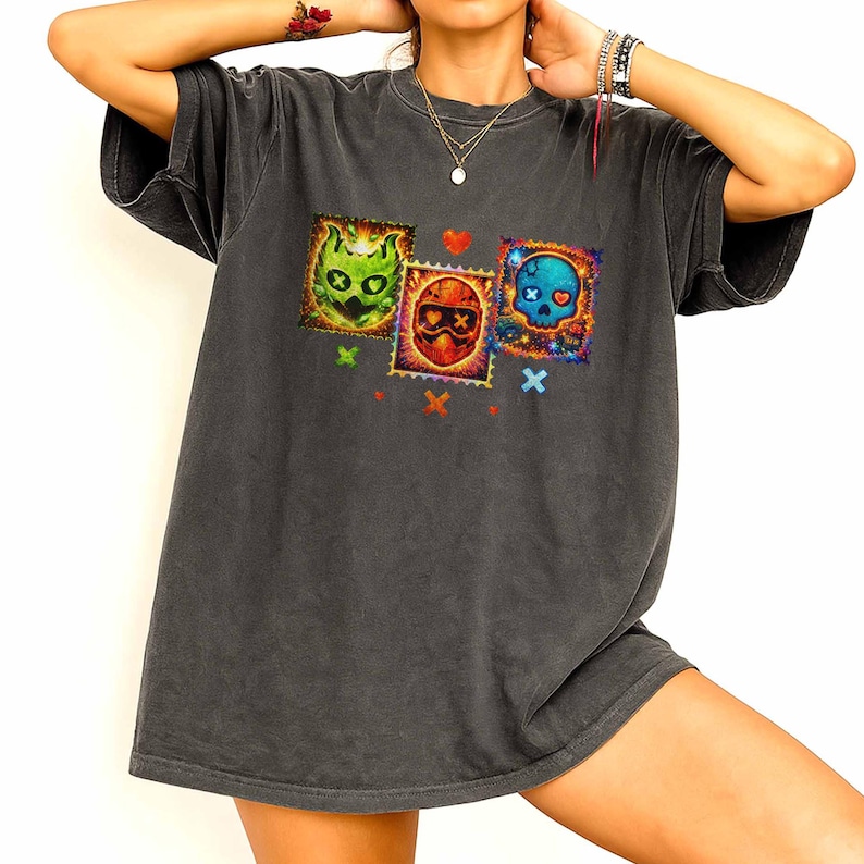 K&ouml;nnte beinhalten: Dunkelgraues T-Shirt mit drei farbenfrohen Totenkopf-Briefmarken-Designs. Die Briefmarken zeigen einen gr&uuml;nen Totenkopf, einen roten Totenkopf und einen blauen Totenkopf, jeweils mit einzigartigen Details und Kreuzstich-Akzenten. Das Shirt hat eine lockere Passform.