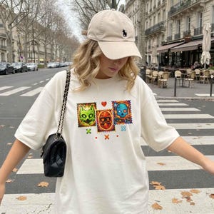 K&ouml;nnte beinhalten: Cremefarbenes T-Shirt mit drei farbenfrohen, stempelartigen Designs mit Monster- und Sch&auml;delgrafiken. Eine beige Baseballkappe mit schwarzem Logo und eine schwarze Umh&auml;ngetasche vervollst&auml;ndigen den Look. Die Person steht auf einem Zebrastreifen.