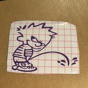 May include: A purple vinyl decal sticker featuring a cartoon character with spiky hair wearing a striped shirt and trainers, depicted in a classic urinating pose. The design is printed on a white backing paper with a red grid pattern overlay.