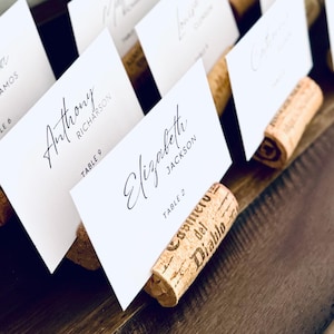 May include: White place cards with elegant calligraphy names and table numbers are displayed in a wooden holder. The cards are held in place by repurposed corks. The arrangement is set on a dark wooden surface, creating a rustic and sophisticated look.