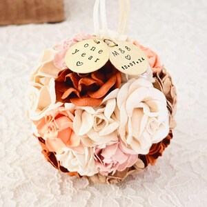 An elegant paper flower ornament in shades of ivory, peach, blush and terra cotta rests on an ivory lace surface.  This ornament was made echoing the color palette from the wedding bouquet in the previous image.  Personalized charms on the ornament read "one year" anniversary, and a couples' initials and wedding date.  The image suggests an elegant, custom first anniversary gift.