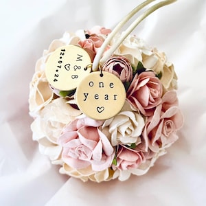 A round, ball-shaped paper flower ornament to celebrate the 1st wedding anniversary rests on pure white tissue paper, suggesting an elegant one year anniversary gift.  The flowers are handmade of paper, which is the traditional gift theme for the first year of marriage.  The paper flowers are very detailed roses and peonies.  The flowers are blush pink, white, ivory, and white.  The display suggests a beautiful keepsake first anniversary gift.
