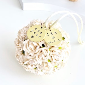 A white flower ornament with 2 personalized engraved charms to celebrate a couples' first wedding anniversary.  One charm reads "one year" with a small heart, the other is J & S with a small heart and wedding date.  The charms are pure brass.  There is a white satin string attached to hang the ornament.