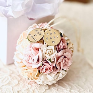 The one year paper flower ornament is shown in front of a glossy white gift box and white tissue paper, both included with your order.  The ornament also includes two brass personalized charms, engraved with "one year" and a couples' initials and wedding date.  The paper flowers are very detailed roses and peonies, in shades of white, ivory, and blush pink.