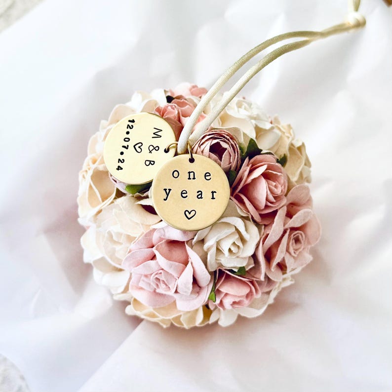 A round, ball-shaped paper flower ornament to celebrate the 1st wedding anniversary rests on pure white tissue paper, suggesting an elegant one year anniversary gift.  The flowers are handmade of paper, which is the traditional gift theme for the first year of marriage.  The paper flowers are very detailed roses and peonies.  The flowers are blush pink, white, ivory, and white.  The display suggests a beautiful keepsake first anniversary gift.