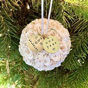First anniversary ornament with two gold charms on a white flower ball. One charm says 'C&M' with a heart and the date '11.19.22'. The other charm says 'one year' with a heart.  The ornament features a simple, elegant design, perfect for commemorating a first wedding anniversary.
