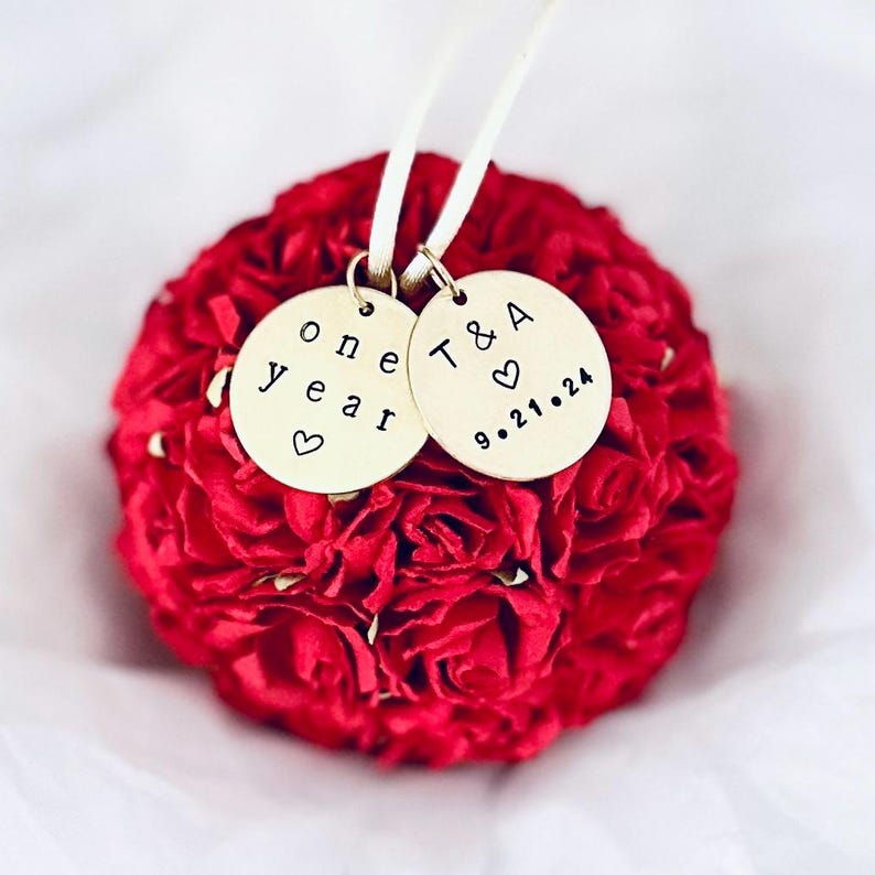A red paper flower Christmas ornament celebrates a one year wedding anniversary.  The flowers are made of paper, with very detailed and realistic petals and tiny green leaves.  There are 2 personalized engraved charms attached to the ornament.  One charm reads "one year" and the second charms reads T & A with wedding date 9.21.24.
