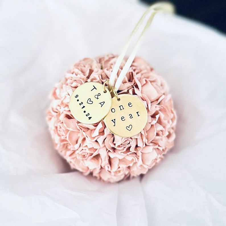 a blush pink flower ornament to celebrate a one year anniversary.  The ball shaped ornament is made of over 40 paper flowers, a nod to the traditional theme of paper for a 1st anniversary gift.  The ornament has two personalized charms attached to the satin hanger.  One charm reads "one year" with a small heart.  The other charm is the initials T & A with anniversary date.