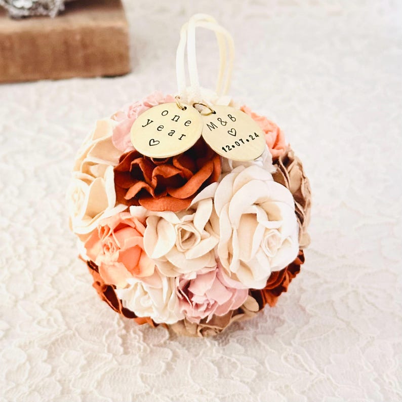 An elegant paper flower ornament in shades of ivory, peach, blush and terra cotta rests on an ivory lace surface.  This ornament was made echoing the color palette from the wedding bouquet in the previous image.  Personalized charms on the ornament read "one year" anniversary, and a couples' initials and wedding date.  The image suggests an elegant, custom first anniversary gift.