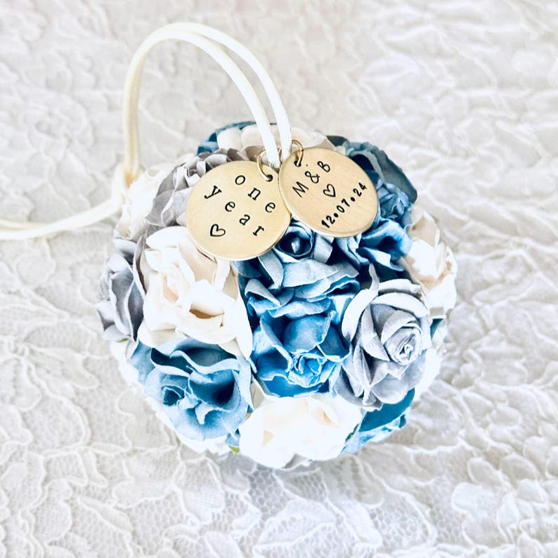 ball shaped ornament made from blue, white and silver paper flowers.  There are personalized charms attached to an ivory satin hanger.  The charms read "one year" for a 1st anniversary gift, and a couples' initials and wedding date.