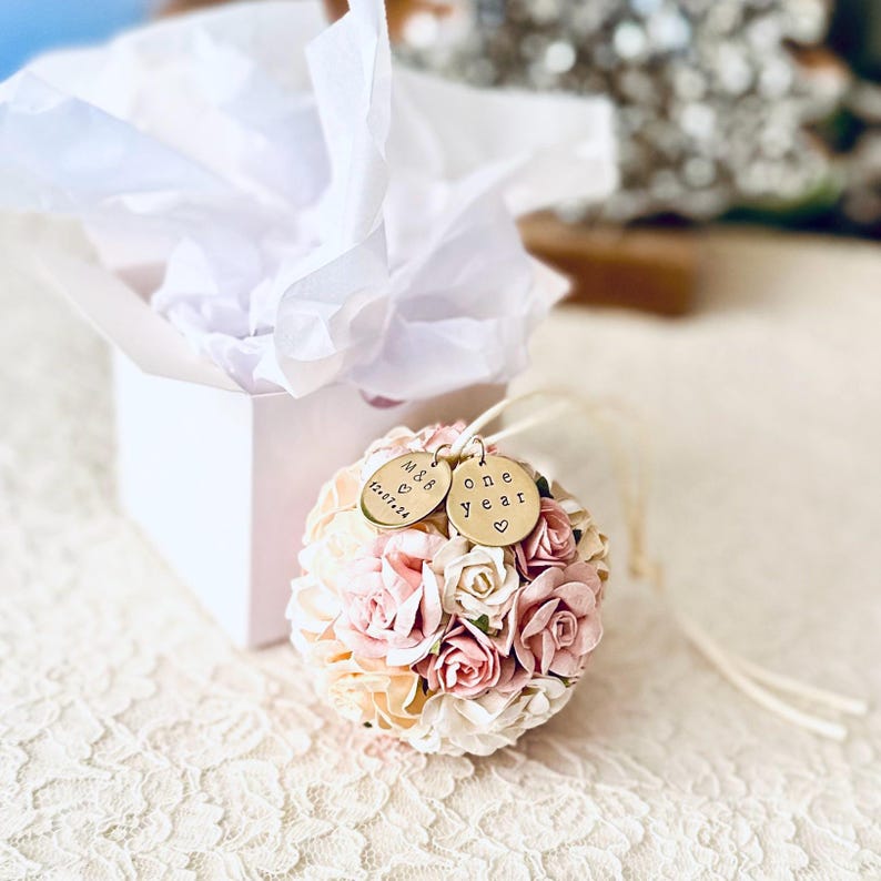 The one year paper flower ornament is shown in front of a glossy white gift box and white tissue paper, both included with your order.  The ornament also includes two brass personalized charms, engraved with "one year" and a couples' initials and wedding date.  The paper flowers are very detailed roses and peonies, in shades of white, ivory, and blush pink.