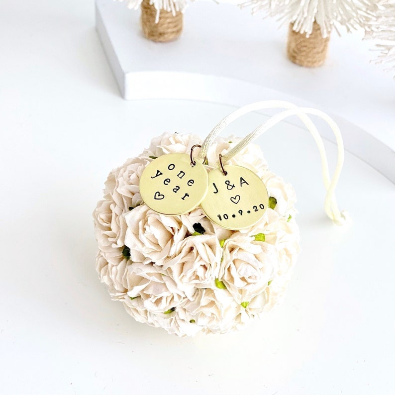 A white flower ornament with 2 personalized engraved charms to celebrate a couples' first wedding anniversary.  One charm reads "one year" with a small heart, the other is J & S with a small heart and wedding date.  The charms are pure brass.  There is a white satin string attached to hang the ornament.