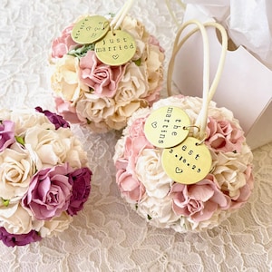 3 custom color paper flower bouquet ornaments are shown in shades of white, ivory, blush pink, and purple.  Each flower ball has 2 personalized brass charms attached to a satin hanger string.  There is a white gift box with white tissue paper in the background, suggesting a beautiful gift idea to celebrate weddings, engagement, or a special anniversary.