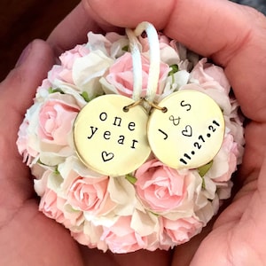 a ball shaped ornament made from blush pink and white paper flowers.  There are 2 personalized charms attached to the ornament hanger string.  One charm reads "one year" with a small heart.  The 2nd charm reads "J & S" with a small heart and wedding date 11.27.21.  This ornament is to celebrate a 1st wedding anniversary.