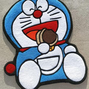 May include: A blue, white, and red cartoon character rug. The character is smiling and holding a brown cookie. The rug has a black border and is on a neutral-colored surface. The rug is shaped like the cartoon character.