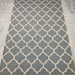May include: A rectangular area rug with a repeating geometric pattern. The rug features a light gray background with a cream-colored quatrefoil design. The rug is made of a woven material and is displayed on a marble floor.