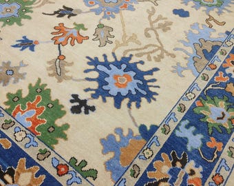 May include: A beige rug with a detailed floral pattern. It has a dark blue border with floral designs in orange, green, and blue. The central design features blue and orange floral motifs.