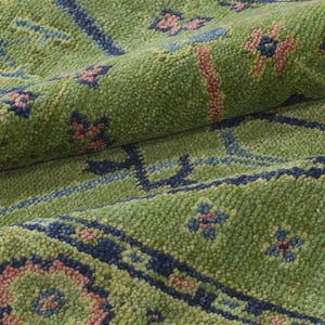 May include: Close-up of a green rug with a textured surface. The rug features a repeating pattern of blue and pink floral and geometric designs. The rug is folded, showing the texture and depth of the material.