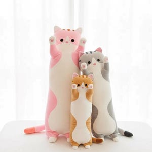 Może przedstawiać: Trzy pluszowe zabawki w kształcie kota w rzędzie. Największy jest różowy i biały, środkowy pomarańczowy i biały, a ostatni szary i biały. Wszystkie trzy mają łapy w górze. Zabawki są miękkie i przytulne.